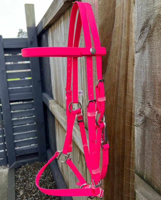 A vibrant LS Deux Bridle in Hot Pink by LS Equestrian hangs on a wooden fence, highlighting its sturdy metal buckles and fittings. The scene is beautifully set against a grey wooden fence and rich greenery in the background.